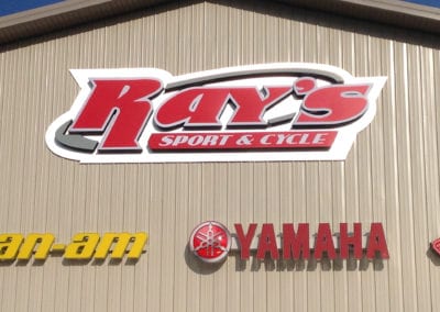 lighted signs on a medal buliding. The Sign is red, gray and white and advertises Ray's Sport & Cycle.